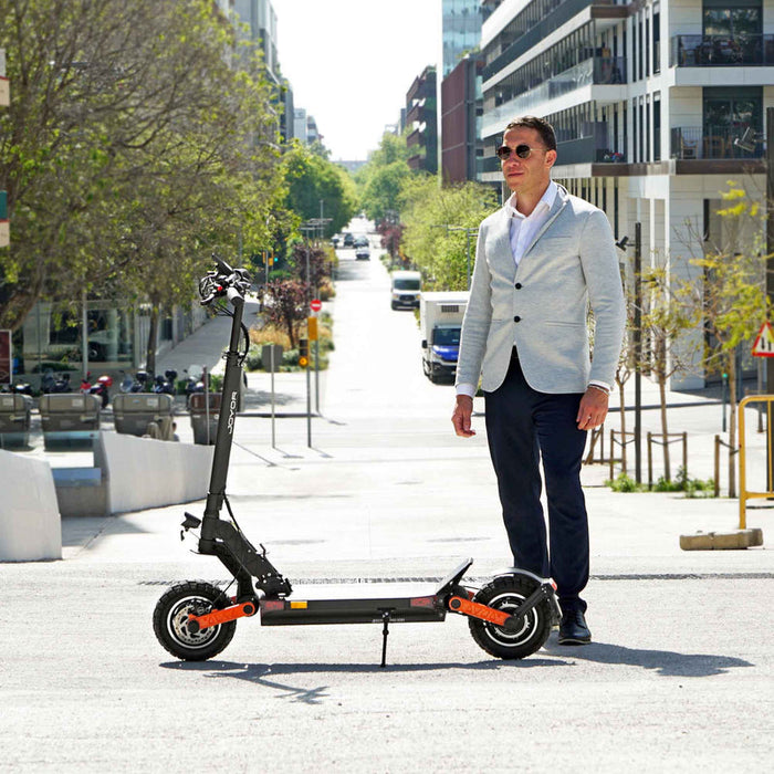 Ein Mann steht neben einem Elektro-Scooter auf einer Straße. Er trägt einen Anzug und hält ein Smartphone in der Hand.
