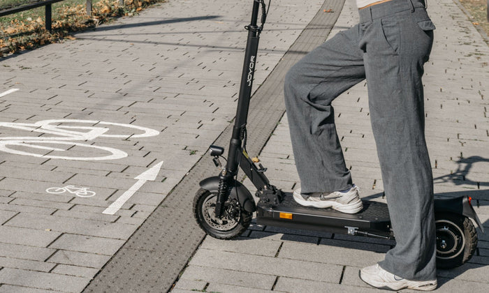 Eine Person in grauer Hose und weißen Schuhen steht auf einem schwarzen Elektroroller auf einem Radweg.