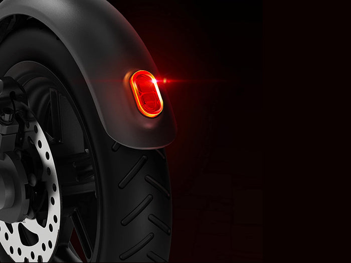 Eine Nahaufnahme des Hinterrads eines Motorrollers mit einem hellroten Rücklicht, das vor einem dunklen Hintergrund leuchtet.