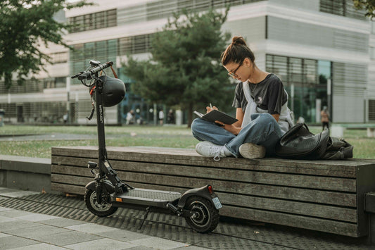 Eine Frau sitzt auf einer Bank, schreibt in ein Notizbuch. Neben ihr steht ein Elektro-Scooter.