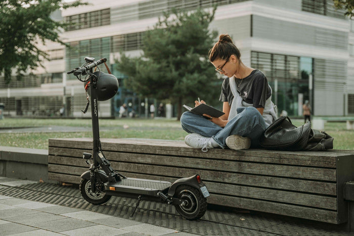 Eine Frau sitzt auf einer Bank, schreibt in ein Notizbuch. Neben ihr steht ein Elektro-Scooter.
