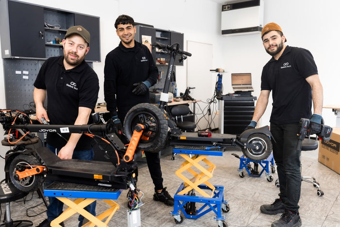 Drei Männer in schwarzer Arbeitskleidung stehen in einer Werkstatt. Sie arbeiten an E-Scootern und anderen Geräten.