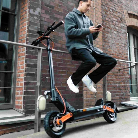 Ein Mann sitzt auf einem Elektro-Scooter vor einer Backsteinmauer und schaut auf sein Smartphone. Er trägt eine Jacke und Sneaker.