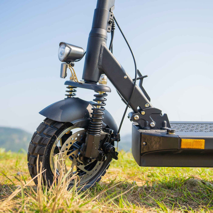 Ein Elektro-Scooter steht auf Gras. Detailansicht des Vorderrades, Federung und Scheinwerfer. Hintergrund: blauer Himmel.