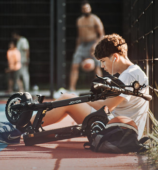  Ein Junge sitzt auf dem Boden neben einem umgestürzten Escooter. Im Hintergrund spielen Menschen Basketball.
